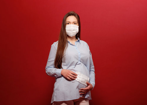 coronavirus pandemic, young pregnant woman on red background in protective medical mask holds on stomach