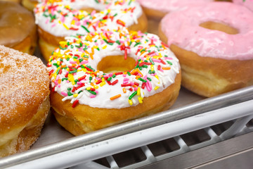 A closeup view of several popular donuts in a bakery display, featuring a glazed donut with rainbow sprinkles.