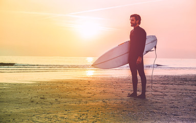 Male surfer standing on the beach waiting for waves at sunset time - Man with surf board wearing wet suit looking the waves - Extreme sport concept - Radial yellow and purple filter editing