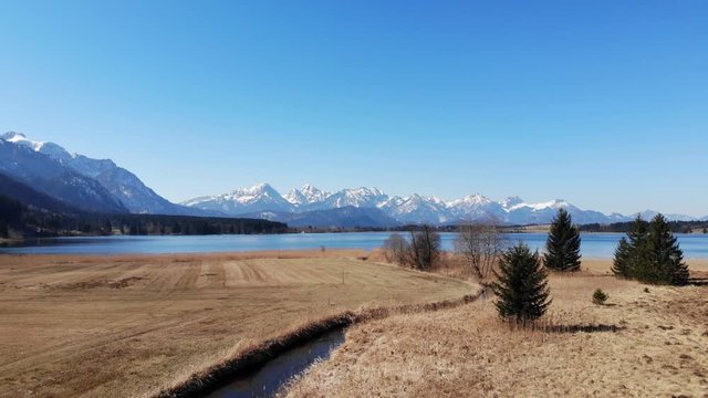 Anflug auf den Bannwaldsee und die Tannheimer Berge mit schneebedeckten Gipfeln im Fr&uuml;hjahr mit Buchinger Bach im Vordergrund