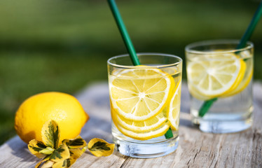 Summer drink, glasses of water filled with slices of lemons, stand on a wooden board on the lawn in sunny warm day, the background is blurred, shallow depth of field, selective focus.