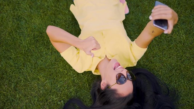 Happy laughing latino brunette girl lying on the grass and taking a selfie. 4K