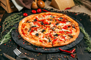 Delicious pizza with mushrooms, olives and pepper on the black board on the dark wooden table with herbs and tomatoes, horizontal, side view