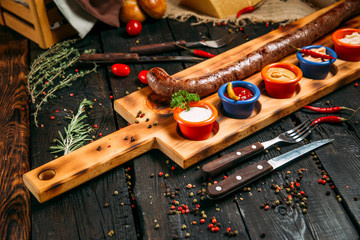 Grilled ground beef sausage with different sauces on a wooden board on a dark wooden background, horizontal