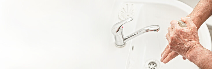 Senior elderly man his hands with soap bar under tap water faucet, wide banner empty space for text left. Can be used as hygiene illustration concept during coronavirus / covid-19 outbreak prevention