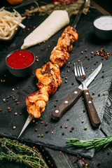 appetizing chicken kebab stick with red sauce on a black board on a dark wooden background, side view, vertical
