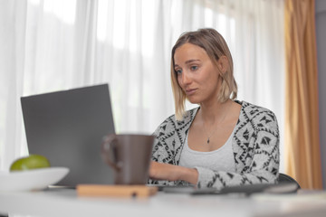 Beautiful girl working from home with laptop sitting at the white table in the bright room with notebook, teacup  and smartphone on the table near, looking at the monitor 