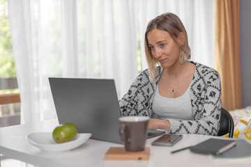 Beautiful girl working from home with laptop sitting at the table in the bright room with notebook,...