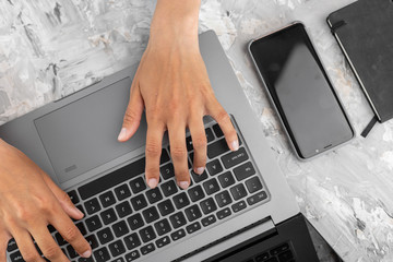 Young woman's hands on the keyboard of the laptop, typing, smartphone and notebook near, abstract grey background, top view, flat lay