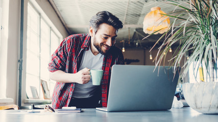 Smiling man working with laptop. Entrepreneur wearing casual clothes and stubble in big spacious bright room. Workspace on the table