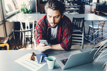 Young man working with laptop. Entrepreneur wearing casual clothes and stubble in big spacious bright room. Workspace on the table. Texting message with mobile phone