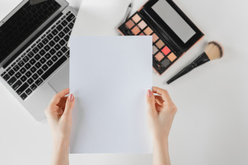Overhead view of female hands holding white paper mockup on white desk with laptop and cosmetics. Flat lay, copy space