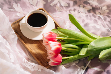 A cup of hot and fragrant freshly ground coffee with a fresh bouquet of white-pink tulips on a wooden tray. Top view