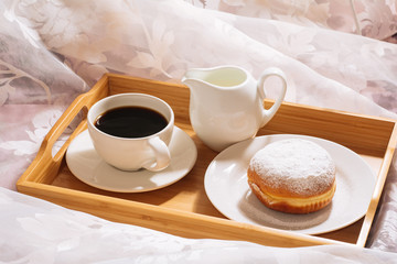 Morning breakfast in bed with fresh coffee, milk and doughnut in powdered sugar, in white dishes on a wooden tray.