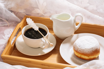 Morning breakfast in bed with freshly brewed coffee, crystal sugar, milk and doughnut in powdered sugar, on a wooden tray in white dishes. Close up