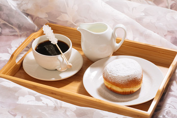 Brunch in bed with freshly roasted and hot bracing coffee, jug of milk, doughnut in powdered sugar on a wooden tray in white dishes