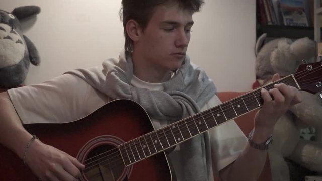 young man wearing casual t-shirt playing acoustic guitar while sitting on sofa in apartment
