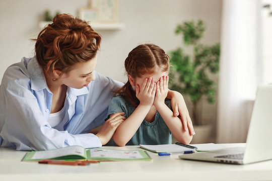 Kind mother embracing and comforting small crying daughter having difficulties with homework while sitting at table and doing exercise in copybook in cozy apartment.