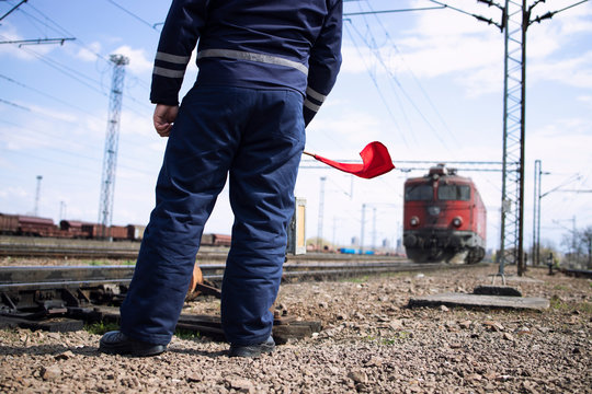 Railroader or switchman at station waving with red flag to the incoming train and signalizing locomotive to slow down or stop.