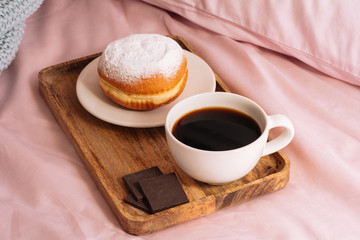 Morning breakfast in bed with fresh and delicious coffee, a doughnut in powdered sugar and three slices of chocolate on a wooden tray against a backdrop of pink sheets