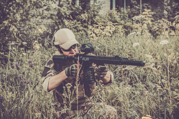 A military man or airsoft player in a camouflage suit sits in the grass and aims from an automatic...