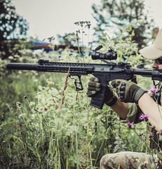 A military man or airsoft player in a camouflage suit sits in the grass and aims from an automatic rifle