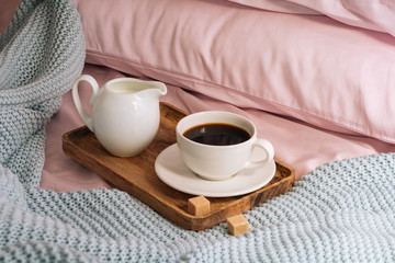 Breakfast in bed with fresh, aromatic and delicious coffee, a jug of cold milk and cane sugar on a wooden tray against a backdrop of pink sheets, pillows and a blue plaid