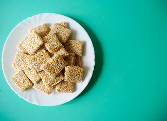 In a white round plate are square-shaped cookies of white sesame, honey and gluten-free sugar on a turquoise background