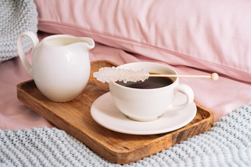 Fototapeta na wymiar Breakfast in bed with freshly brewed and delicious coffee, a jug of cold milk and crystal sugar on a stick on a wooden tray against a backdrop of pink sheets, pillows and a blue plaid