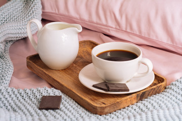 Breakfast in bed with freshly brewed and aromatic coffee, a jug of cold milk and three slices of real dark chocolate on a wooden tray against a backdrop of pink sheets, pillows and a blue plaid