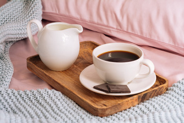 Breakfast in bed with freshly brewed and delicious coffee, a jug of cold milk and three slices of real dark chocolate on a wooden tray against a backdrop of pink sheets, pillows and a blue plaid