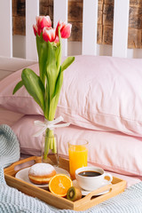 breakfast in bed with a cup of freshly brewed coffee, orange juice, orange, sweet kiwi, doughnut and a bouquet of freshly cut tulips in vase in a wooden tray. background of pink pillows and blue plaid