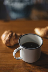 enamelled cup of coffee on wooden table in front of a croissant and a french press