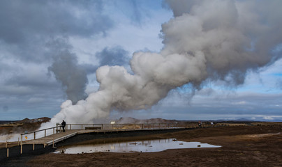 Fototapeta na wymiar Geothermal area in Iceland. Lonely person. Powerful steam jet above the ground. A smoking geyser on a background of yellow clay and a cloudy sky. Reykjanes Peninsula