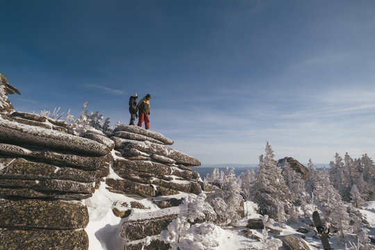 Hikers couple relaxing on top of mountain cliff and enjoying the view of valley and winter mountain landscape. Freeride snowboarding, sunny day