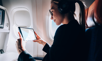 Caucasian woman in bluetooth headphones for noise cancellation choosing media podcast for listening during international jetliner flight, formally dressed female messaging via application on touch pad