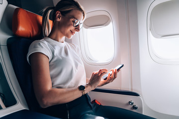 Female tourist in classic eyewear checking received email in social networks during internet messaging on board in jetliner, Caucasian woman booking return tickets connected to high speed wifi
