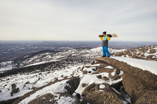 Hiker snowboarder freerider holding snowboard standing on big rock cliff watching mountain landscape