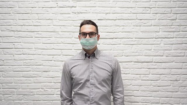 Attractive Caucasian Man Businessman With Brown Hair In Black Glasses In A Checkered Shirt Adjusting And Puts On A Protective Turquoise Medical Mask On His Face Against A White Brick Wall.