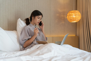 Young asian woman relaxing in the hotel and working with laptop on a bed, calling to cusotmer, Business trip, Working travel concept