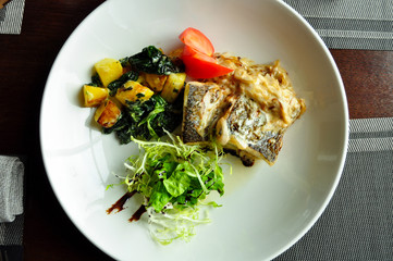 Baked fish in a creamy sauce with baked potatoes, tomatoes and fresh herbs on a white plate. Top view.
