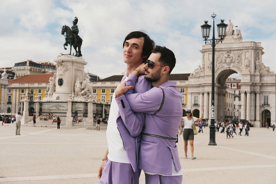 Romantic male couple wearing purple suits travel, walking and posing in European city street. Lisbon, Praca de Comercio