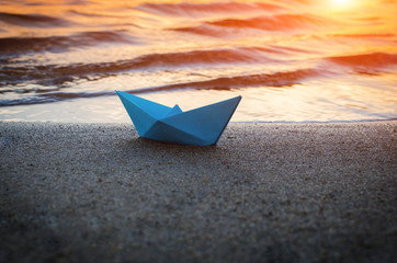 Lone blue paper boat washed up on the sandy beach at sunset.
