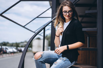 Beautiful girl with long hair and glasses sitting on metal stairs on the wooden background of house with vertical boards. Woman smiling and looking at camera