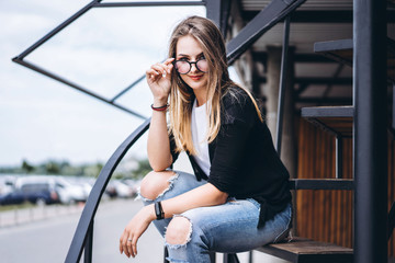 Fototapeta premium Beautiful girl with long hair and glasses sitting on metal stairs on the wooden background of house with vertical boards. Woman smiling and looking at camera