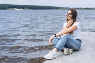 Fototapeta na wymiar Young woman with long hair in stylish glasses posing on the concrete shore near the lake. Girl dressed in jeans and t-shirt smiling and looking away