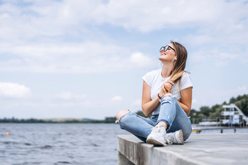 Fototapeta na wymiar Young woman with long hair in stylish glasses posing on the concrete shore near the lake. Girl dressed in jeans and t-shirt smiling and looking away