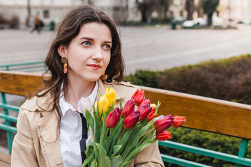 Beautiful young woman with spring tulips flowers bouquet at city street. Happy girl sitting on a bench outdoors. Spring portrait of pretty female in old town