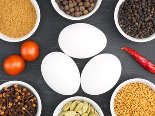 Raw chicken eggs, black pepper, masala powder, fenugreek, green cardamom, hot red chili pepper, cloves, tomato on black wooden background