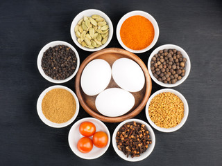 Raw chicken eggs, black pepper, masala powder, fenugreek, green cardamom, hot red chili pepper, cloves, tomato on black wooden background